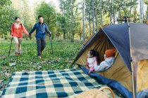 Entzückende Kinder, die im Zelt sitzen und lächelnde Eltern, die mit Trekkingstöcken im Wald spazieren gehen — Stockfoto
