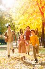 Felices padres jóvenes y lindos niños corriendo en el bosque de otoño y sonriendo a la cámara - foto de stock