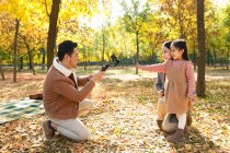 Happy asian father taking photo of daughter and wife with smartphone in autumnal park — Stock Photo