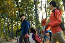 Seitenansicht einer glücklichen jungen asiatischen Familie mit Rucksäcken und Trekkingstöcken, die gemeinsam im herbstlichen Wald spazieren gehen — Stockfoto