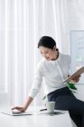 Attraktive asiatische Geschäftsfrau sitzt auf dem Tisch und benutzt Laptop im hellen Büro — Stockfoto