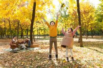 Enfants heureux jouant avec les feuilles d'automne tandis que les parents reposant sur carreaux à carreaux dans le parc — Photo de stock