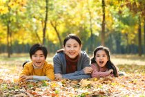 Sorrindo asiática mãe com filha e filho deitado na folhagem no parque outonal e olhando para a câmera — Fotografia de Stock