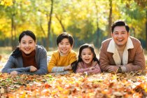 Padres jóvenes felices con dos niños acostados juntos y sonriendo a la cámara en el parque de otoño - foto de stock