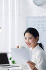 Attraktive lächelnde asiatische Geschäftsfrau trinkt Kaffee und liest Dokumente im hellen Büro — Stockfoto