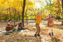 Bambini felici che giocano con le foglie autunnali mentre i genitori riposano sul plaid a scacchi nel parco — Foto stock