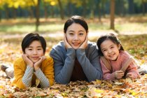Sonriente asiático madre con hija e hijo acostado en follaje en otoñal parque y mirando a cámara - foto de stock