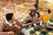 Вид улыбающегося отца, играющего на гитаре, и счастливой семьи, сидящей на чучеле в парке — стоковое фото