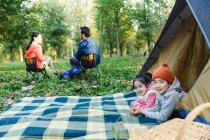 Happy siblings lying in tent and smiling at camera while parents resting behind in forest — Stock Photo