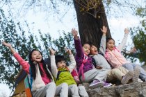 Cinq adorables heureux asiatiques enfants assis sur des pierres avec les mains vers le haut dans le parc automnal — Photo de stock