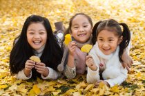 Три чарівні усміхнені азіатські діти лежать на жовтому листі і тримають листя в автономному парку — стокове фото