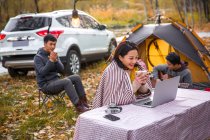 Smiling asian woman holding cup of tea and looking at laptop near camping place in autumnal forest — Stock Photo