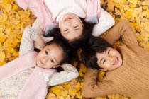 Vista superior de niño feliz y niñas acostados juntos en hojas amarillas y sonriendo a la cámara en el parque de otoño - foto de stock