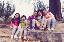 Cinq adorables enfants asiatiques assis sur des pierres et regardant la caméra dans le parc automnal — Photo de stock