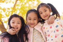 Низький кут зору трьох чарівних усміхнених азіатських дітей, що обіймаються в автономному парку і дивляться на камеру — стокове фото