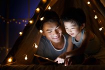 Щасливий азіатський батько і дошкільник син дивиться щось на планшеті в наметі вдома — стокове фото