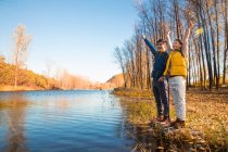 Happy asian couple standing with hands up near lake in autumnal forest — Stock Photo