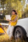 Cheerful asian woman holding laptop and leaning on car in autumnal forest — Stock Photo