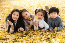 Ragazzo felice e ragazze sdraiate insieme su foglie gialle e sorridenti alla macchina fotografica nel parco autunnale — Foto stock