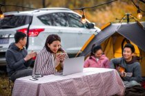 Sorrindo asiático mulher segurando xícara de chá e olhando para laptop perto de camping lugar no outonal floresta — Fotografia de Stock