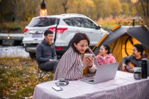 Mulher asiática segurando xícara de chá e olhando para laptop perto de camping lugar na floresta outonal — Fotografia de Stock