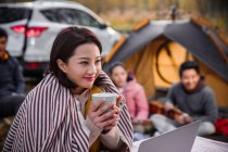 Attractive asian woman holding cup of tea and looking at laptop near camping place in autumnal forest — Stock Photo