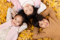 Vista superior de menino feliz e meninas deitadas juntas em folhas amarelas e sorrindo para a câmera no parque de outono — Fotografia de Stock