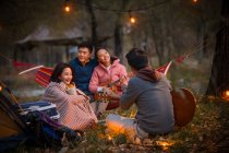 Mann spielt Gitarre für drei asiatische Freunde beim Zelten im herbstlichen Abendwald — Stockfoto
