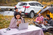 Asian woman holding cup of tea and looking at laptop near camping place in autumnal forest — Stock Photo