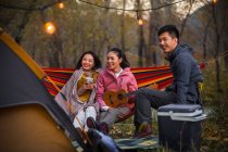 Smiling asian friends having fun with ukulele on hammock in autumnal forest — Stock Photo