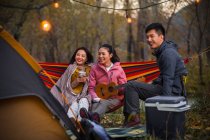 Souriant asiatique amis avoir amusant avec ukulele sur hamac en automne forêt — Photo de stock