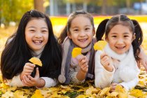 Three adorable happy asian kids lying on yellow foliage and holding leaves in autumnal park — Stock Photo