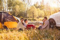 Four happy asian friends drinking tea and talking at camping place in autumnal forest — Stock Photo