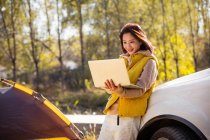 Веселая азиатка, держащая в руках ноутбук и опираясь на автомобиль в осеннем лесу — стоковое фото