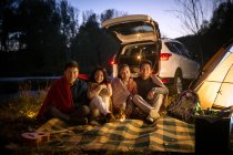 Четыре улыбающихся азиатских друга сидят рядом с машиной и смотрят в камеру в осеннем вечернем лесу — стоковое фото