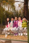 Five adorable smiling asian kids sitting on fence and looking at camera in autumnal park — Stock Photo