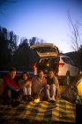 Четыре улыбающихся азиатских друга сидят рядом с машиной и смотрят в камеру в осеннем вечернем лесу — стоковое фото