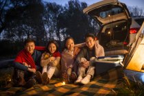 Cuatro sonrientes amigos asiáticos sentados cerca de coche y mirando a la cámara en otoño bosque de noche - foto de stock