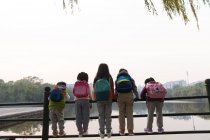 Visão traseira de cinco crianças que se apoiam na cerca perto do rio no parque outonal — Fotografia de Stock