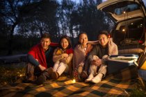 Чотири усміхнені азіатські друзі сидять біля машини і дивляться на камеру в автентичному вечірньому лісі — стокове фото