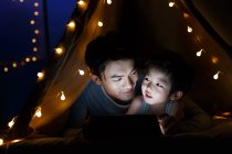 Feliz asiático padre y preescolar hijo viendo algo en tableta en tienda de campaña en casa - foto de stock