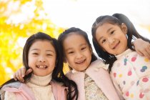 Niedrigwinkel-Ansicht von drei entzückenden lächelnden asiatischen Kindern, die sich im herbstlichen Park umarmen und in die Kamera schauen — Stockfoto