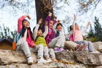 Cinco adorable feliz asiático niños sentado en piedras con manos arriba en otoñal parque - foto de stock