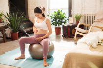 Visão de alto ângulo de sorrir jovem grávida em sportswear sentado na bola em forma em casa — Fotografia de Stock