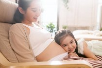 Милая улыбающаяся маленькая девочка слушает живот счастливой беременной матери дома — стоковое фото