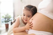 Обрезанный снимок милой улыбающейся дочери, слушающей живот беременной матери — стоковое фото