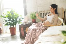 Vista lateral de la joven embarazada feliz sentado en la silla y el uso de teléfono inteligente - foto de stock