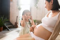 Niedlich lächelnde Tochter hält Stethoskop und hört Bauch der schwangeren Mutter zu Hause zu — Stockfoto