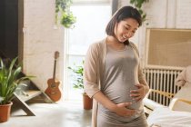 Lächelnde junge asiatische Schwangere, die ihren Bauch berührt und zu Hause nach unten schaut — Stockfoto