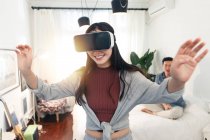 Щаслива молода азіатська жінка використовує гарнітуру віртуальної реальності, а хлопець сидить на ліжку позаду — стокове фото